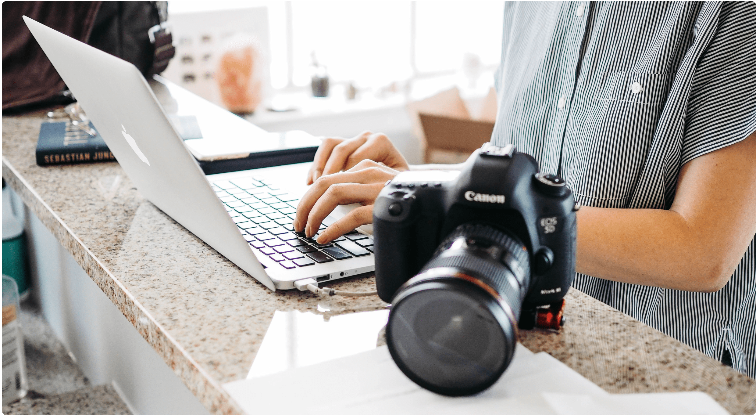 A camera sitting on a counter in front of someone typing on a laptop. Photo from Unsplash.com